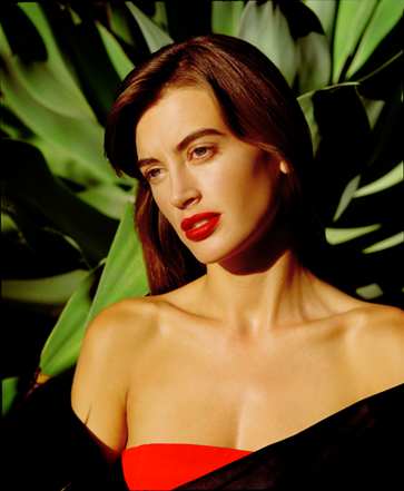 late afternoon sunlit photo of Amanda Pays in Donna Karan red strapless dress with black off the shoulder cape standing in front of lush deep green yucca cactus  fronds in a photograph by celebrity and fashion photographer Steve Landis.